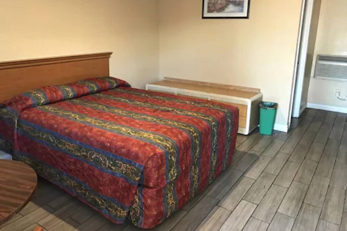 A bedroom with a bed, a wooden headboard, a bench, a picture on the wall, and a trash bin.