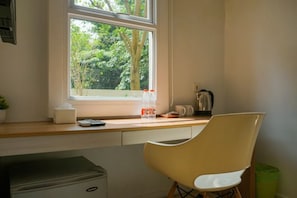 Una cocina con ventana, una silla, un cubo de basura y una cafetera.