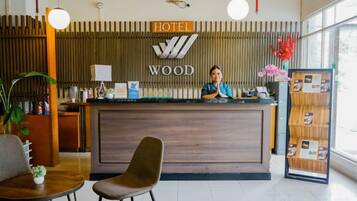Mostrador de recepción de hotel con encimera de madera, una recepcionista y zona de recepción con una planta y un tablón de anuncios.