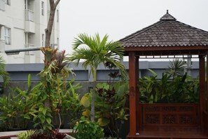 A gazebo with a tiled roof and a wooden structure, surrounded by various plants and a building in the background. - Wood Hotel (Cidadap)