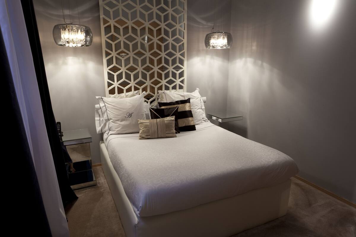 Un lit bien fait, avec du linge de lit blanc et des coussins décoratifs, un mur orné d’un motif géométrique et deux luminaires suspendus.