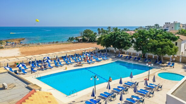A large outdoor swimming pool with blue lounge chairs and umbrellas, surrounded by a sandy beach and palm trees. - Tetyk Hotel  (Paralimni)