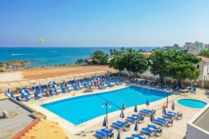 A large outdoor swimming pool with blue lounge chairs and umbrellas, surrounded by a sandy beach and palm trees. - Tetyk Hotel  (Paralimni)