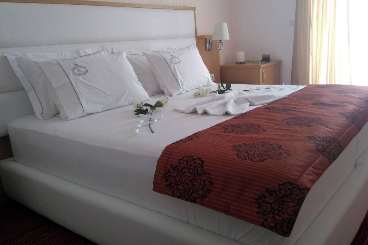 A hotel room with a large bed, white bedding, a red patterned throw, and a nightstand with a lamp.