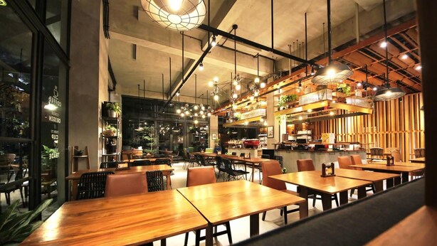 A modern restaurant with wooden tables and chairs, hanging pendant lights, and a visible kitchen area. - Hay Bandung (Bandung)