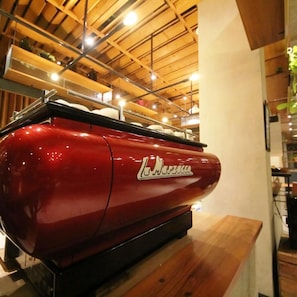 A red barbecue grill with a logo on it, placed on a wooden table in a room with wooden beams on the ceiling. - Hay Bandung (Bandung)