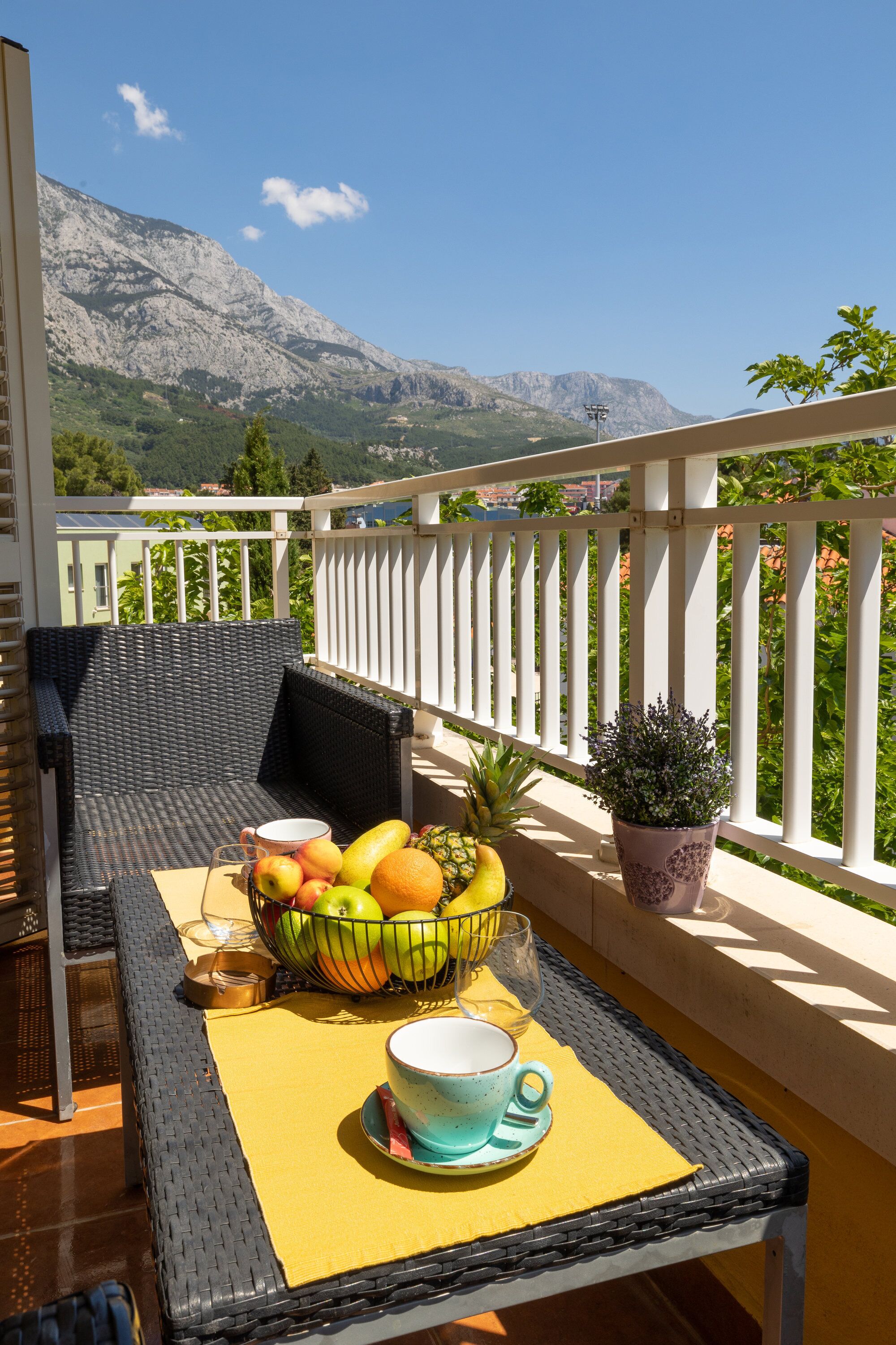 A balcony with wicker furniture, a table with a fruit bowl, a potted plant, and a scenic mountain view.