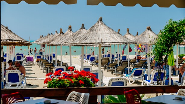 Un restaurante frente a la playa con mesas al aire libre, sombrillas de paja y vistas a la arena.