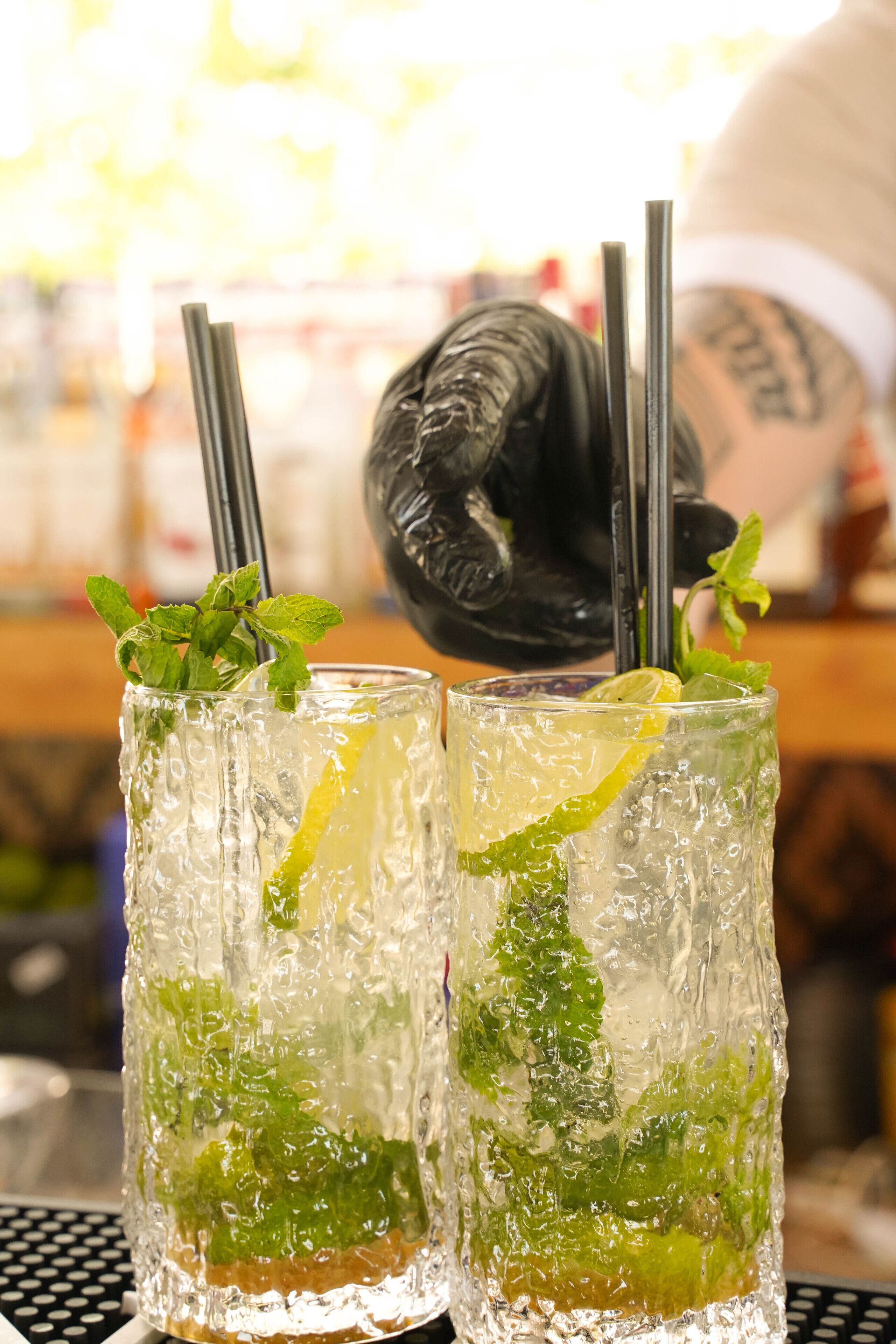 Two tall glasses of a clear beverage with mint and lemon garnish, accompanied by black straws, on a table with a polka dot pattern.