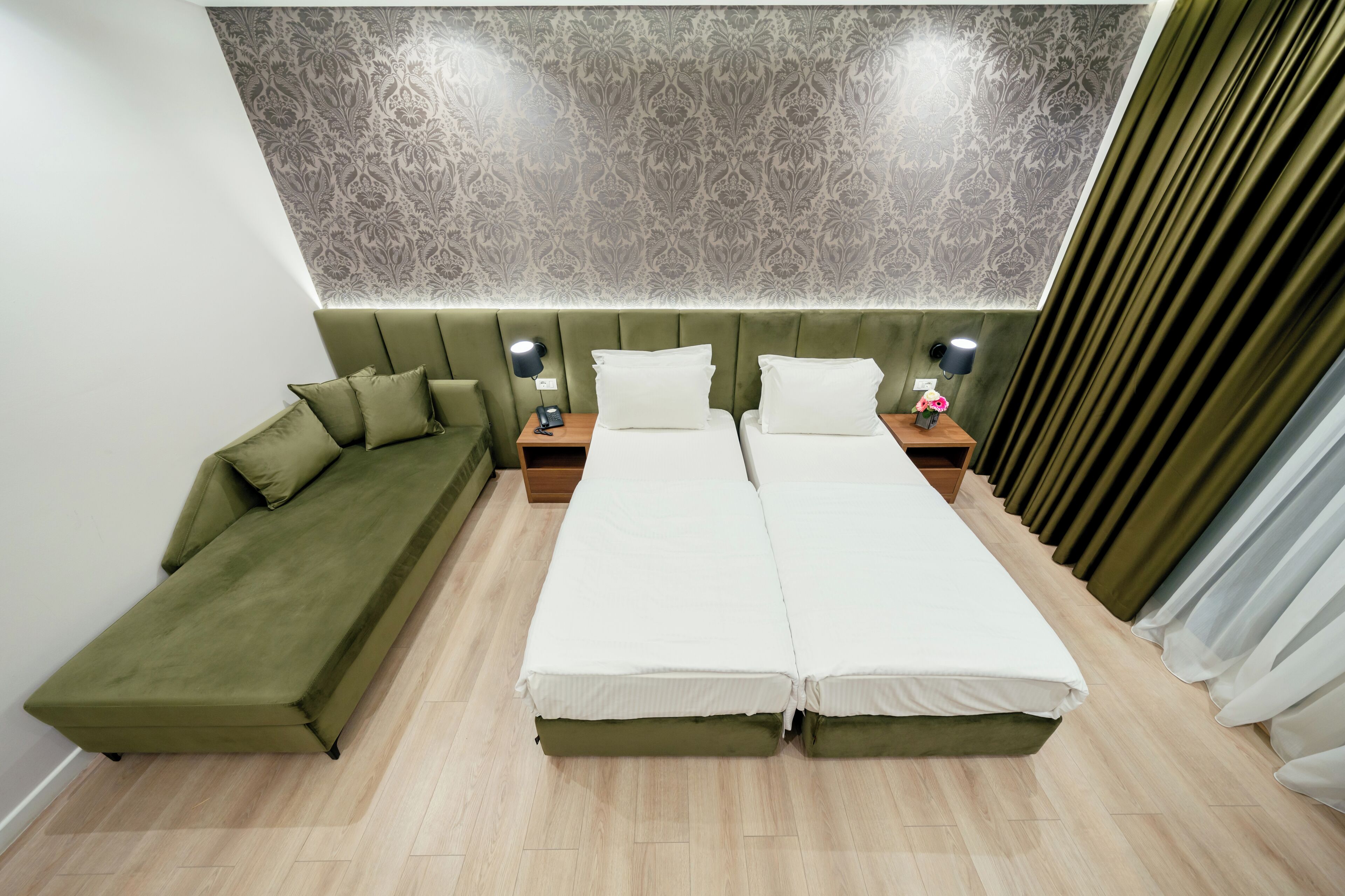 Habitación de hotel con un sofá verde, una cama doble con ropa blanca, una mesita de noche con lámpara y un papel tapiz estampado en la pared.