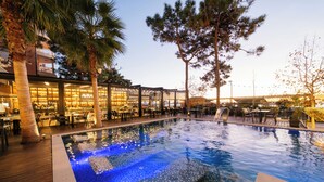 Un área de piscina con solárium de azulejos, sillas de descanso y un restaurante con cerramiento de cristal.