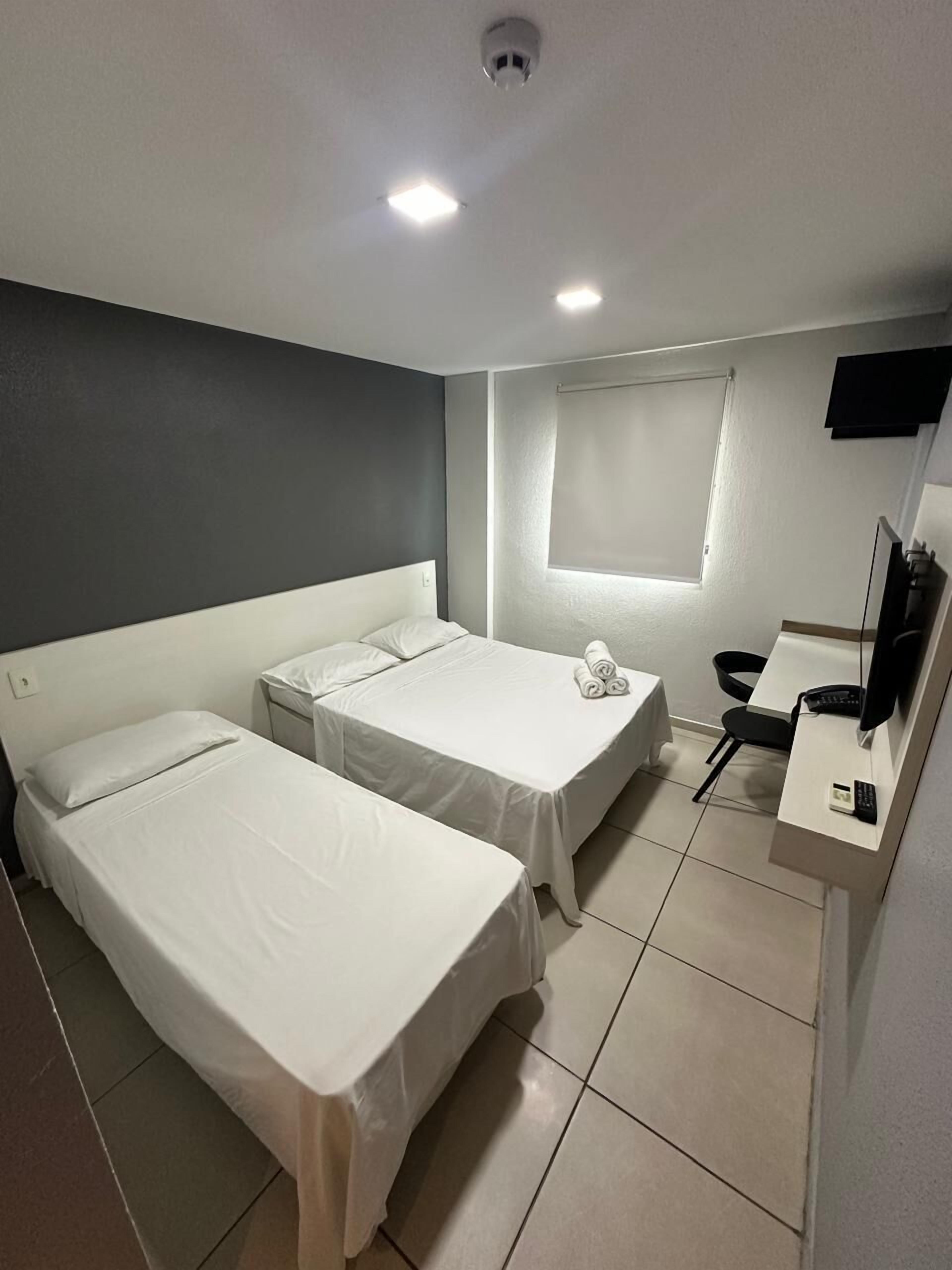 A hotel room with two single beds, a desk with a chair, a TV, and a window with blinds.