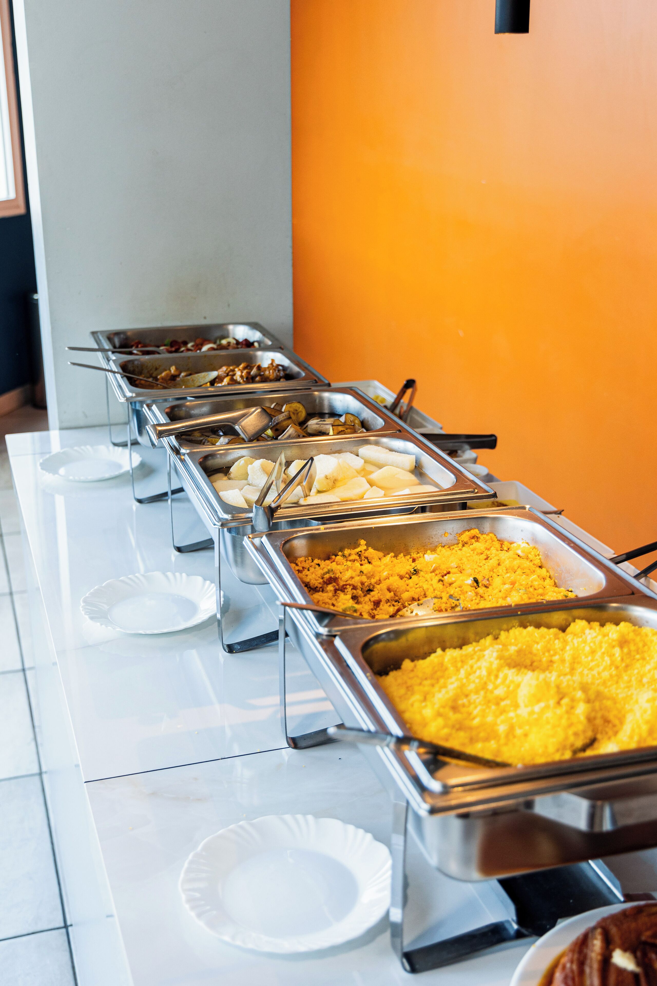 A buffet with various food items in metal containers, white paper plates, and a tiled floor.