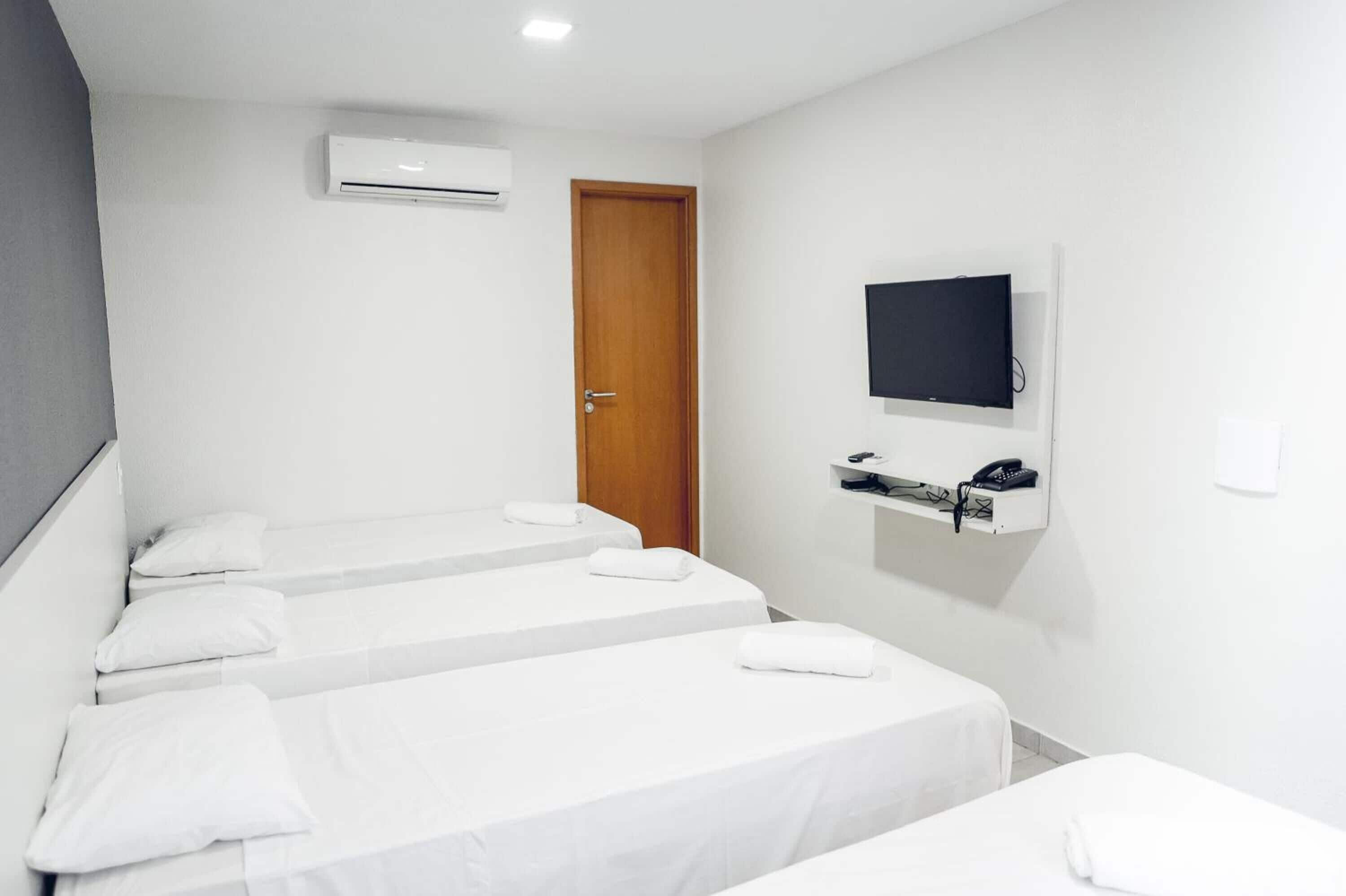 A hotel room with three single beds, a TV mounted on the wall, and a small shelf with a phone and other items.