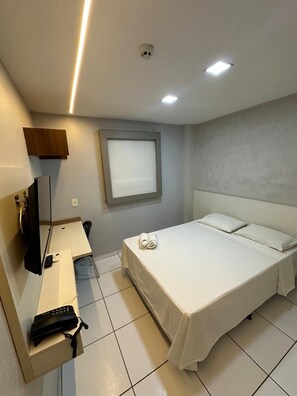 A hotel room with a bed, a desk with a telephone, a television, and a window with blinds.
