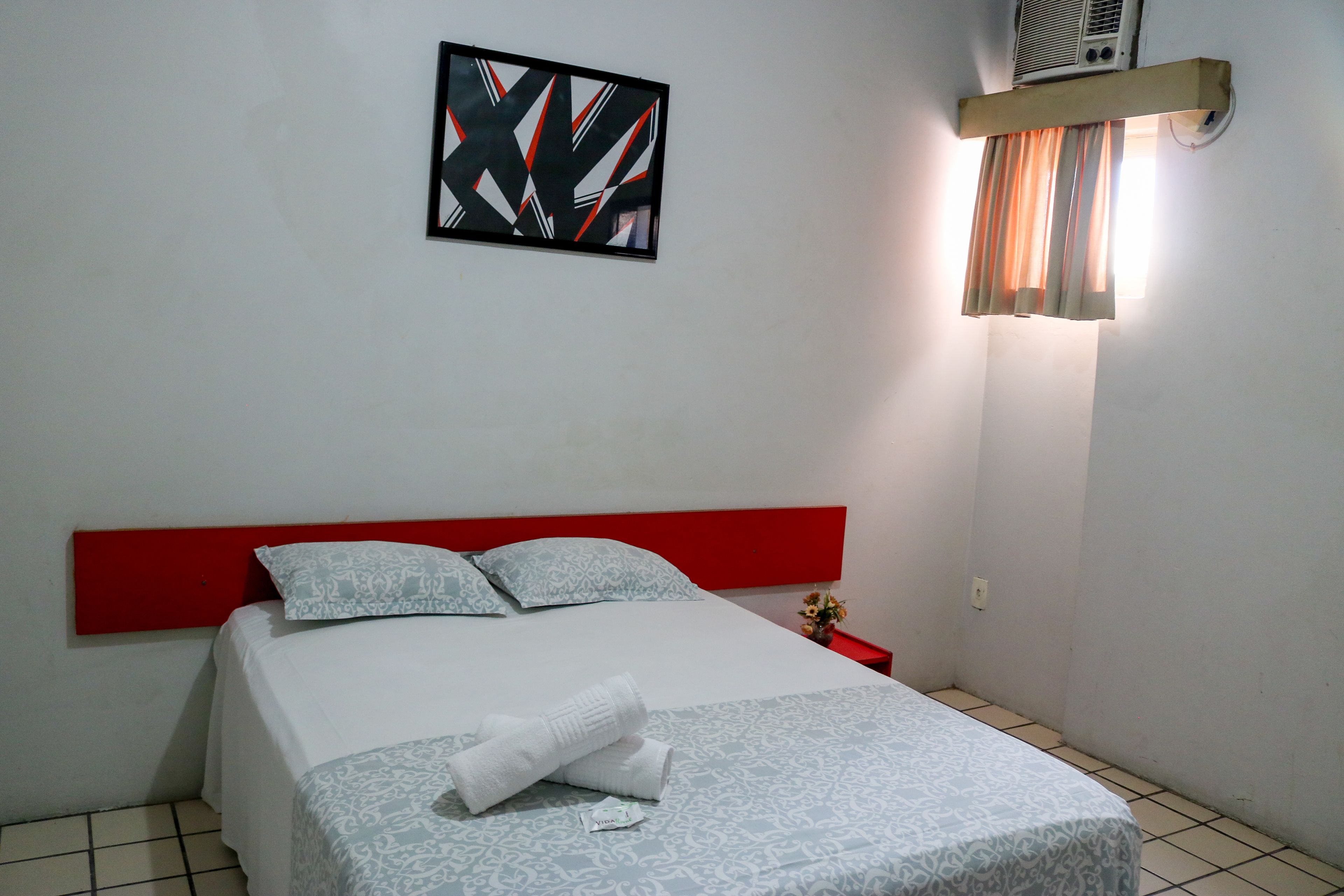 A neatly made bed with a red headboard, a framed abstract artwork on the wall, and a window with curtains.
