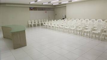 Un salón de conferencias con una hilera larga de sillas de plástico blanco, una mesa verde y luces fluorescentes en el techo.