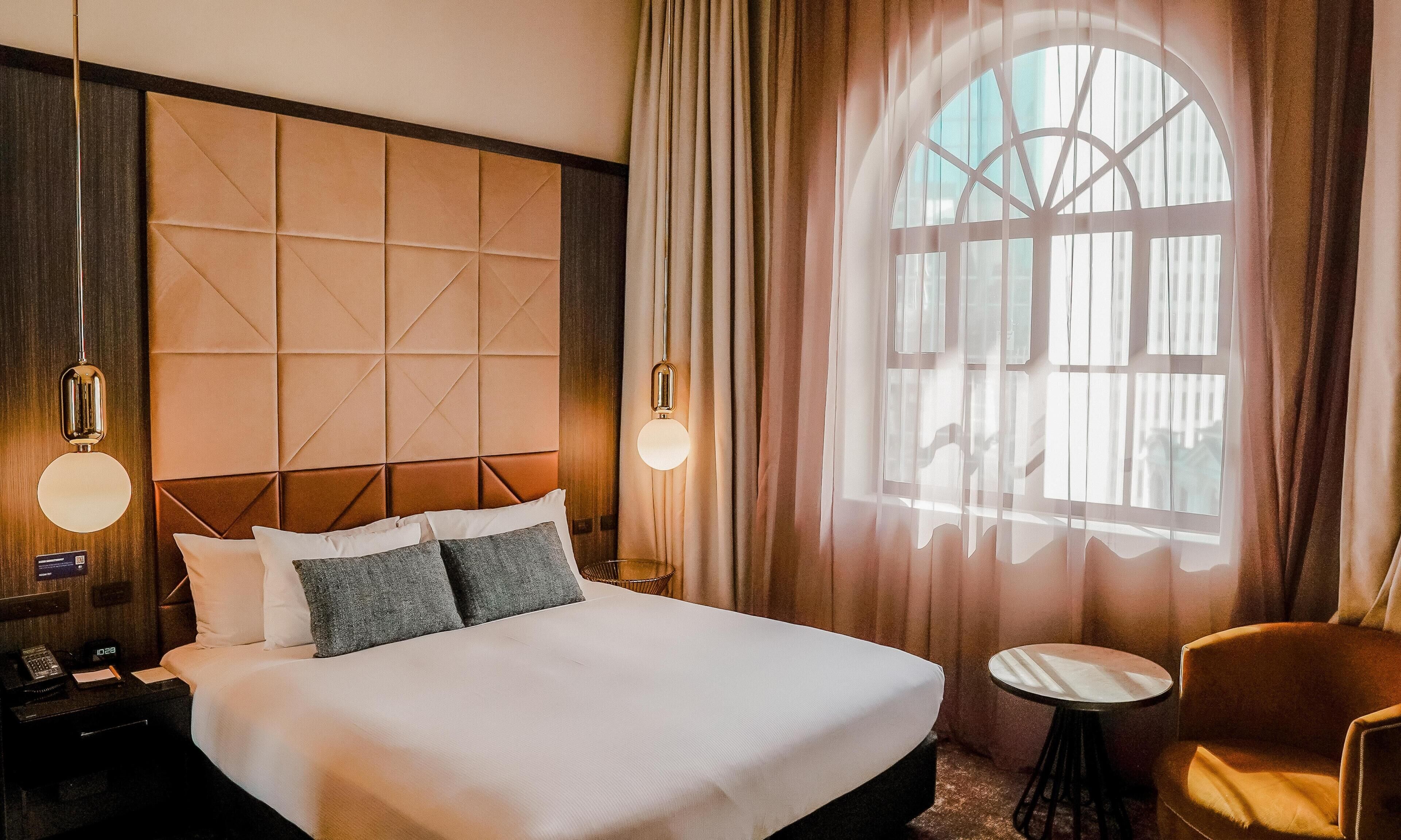 Una habitación de hotel con una cama grande, una mesita de noche con lámpara, una silla y un ventanal con cortinas.