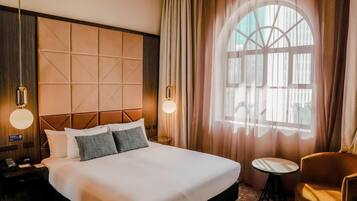 A hotel room with a large bed, a bedside table with a lamp, a chair, and a window with curtains.