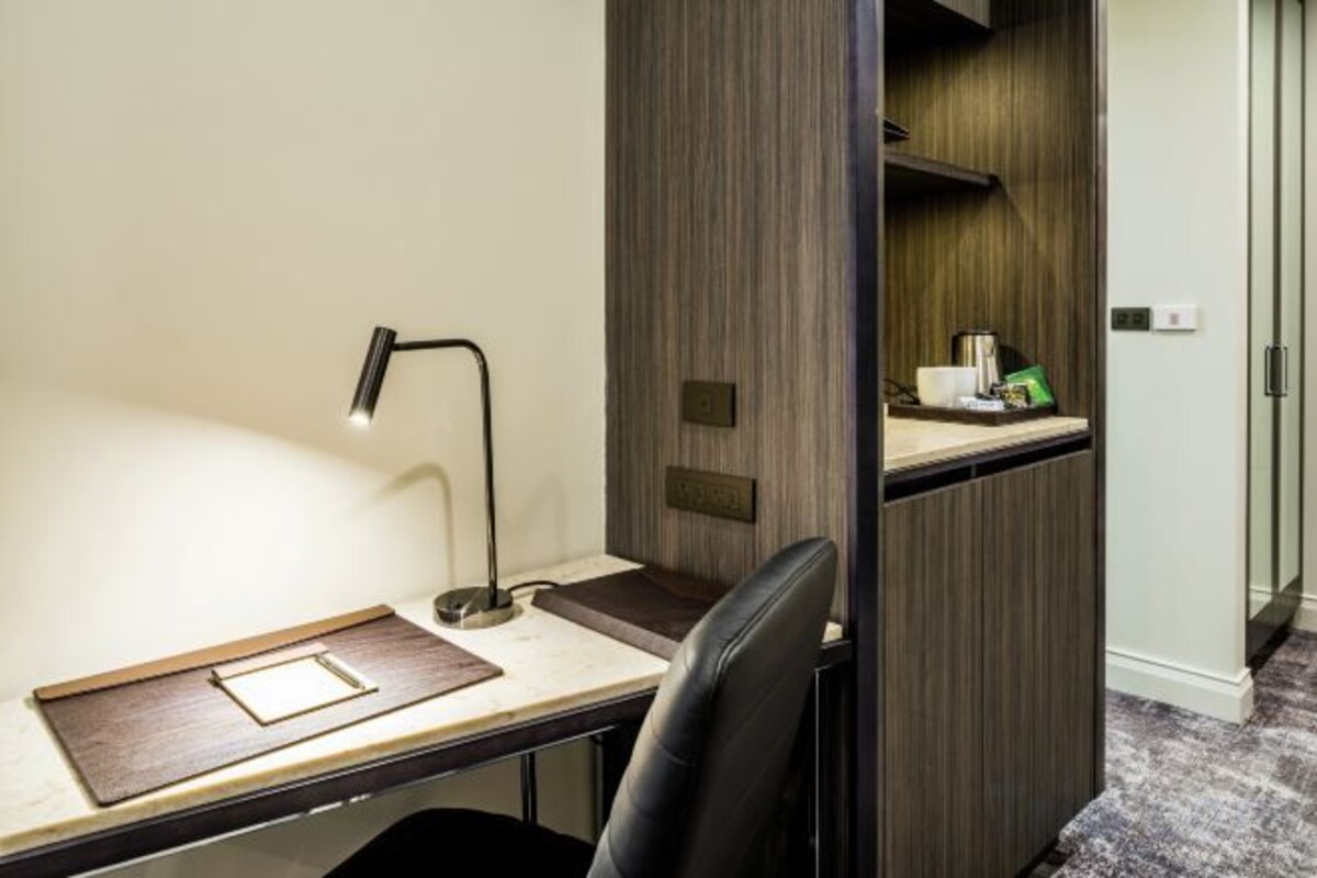 A hotel room with a desk, a chair, a lamp, and a small shelf with toiletries.