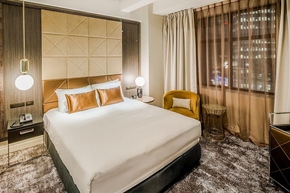 Habitación de hotel con una cama grande, una mesita de noche con lámpara, una silla y vistas a la ciudad a través de la ventana.