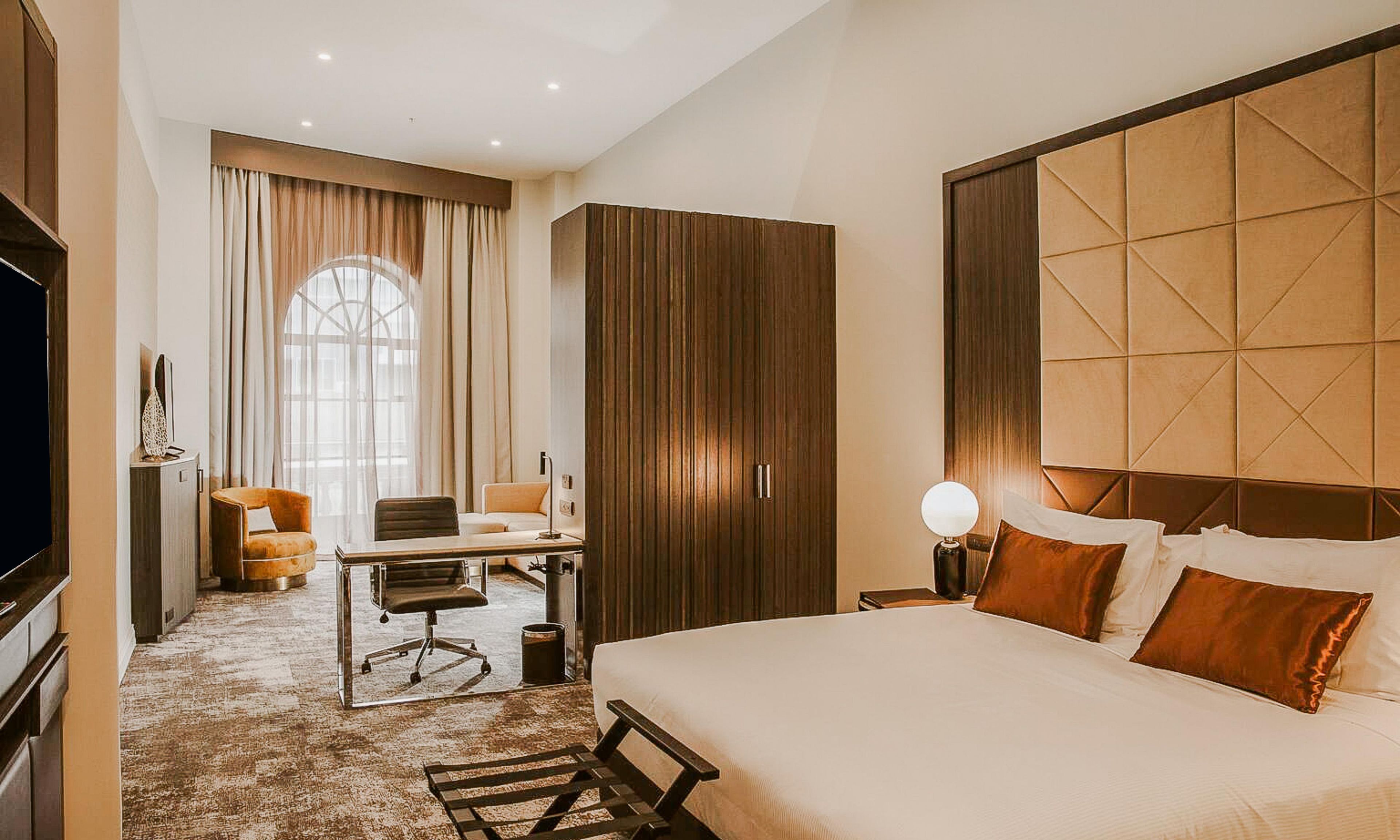 A hotel room with a large bed, a desk with a chair, a wardrobe, and a television.