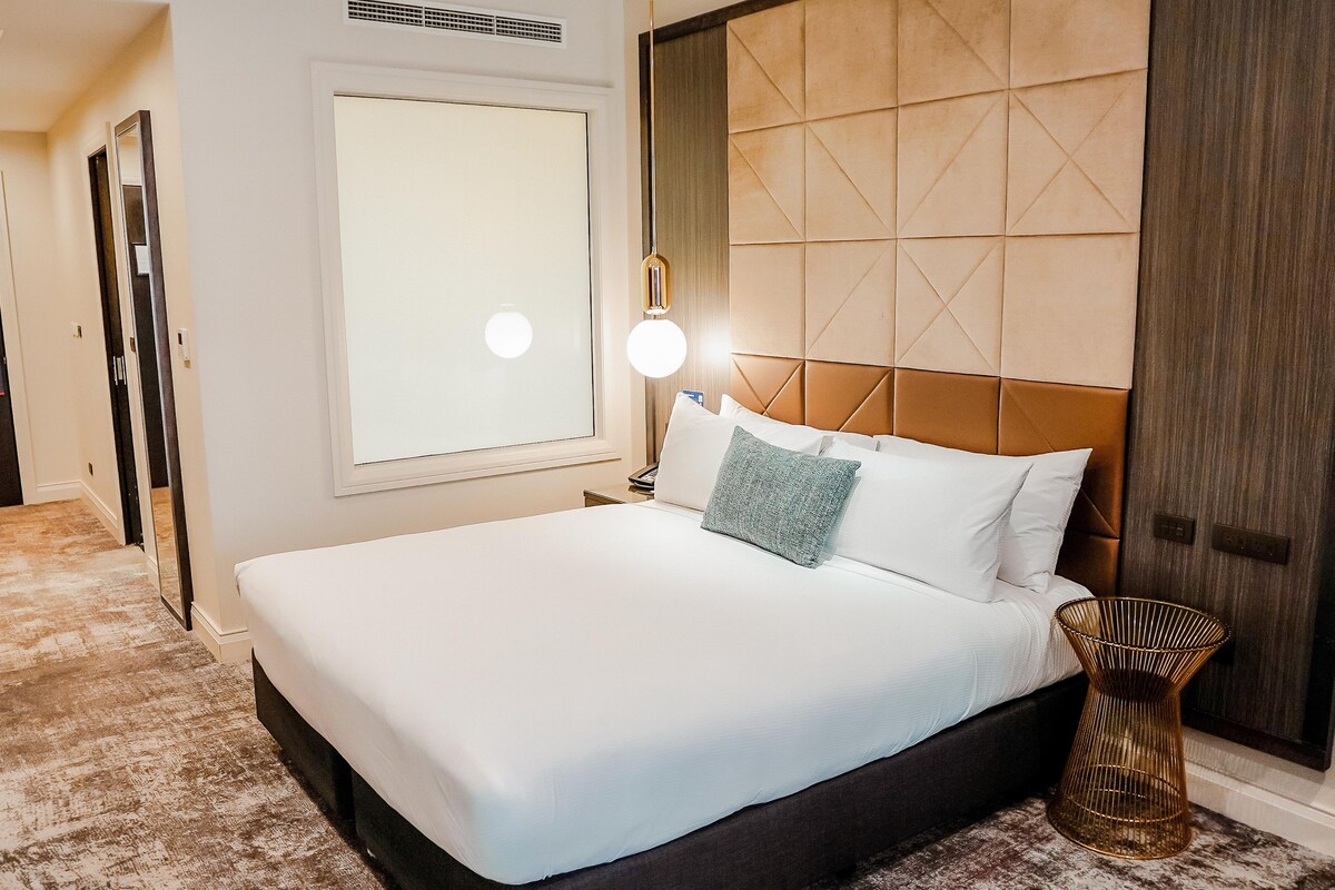 Una habitación de hotel con una cama grande, un televisor montado en la pared, una mesita de noche con lámpara y una puerta con picaporte.