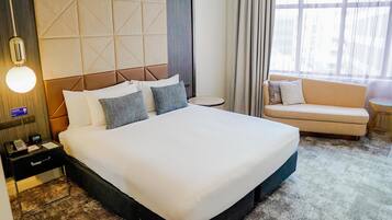 A hotel room with a large bed, a bedside table with a phone and a clock, a small round table, a sofa, and a window with curtains.