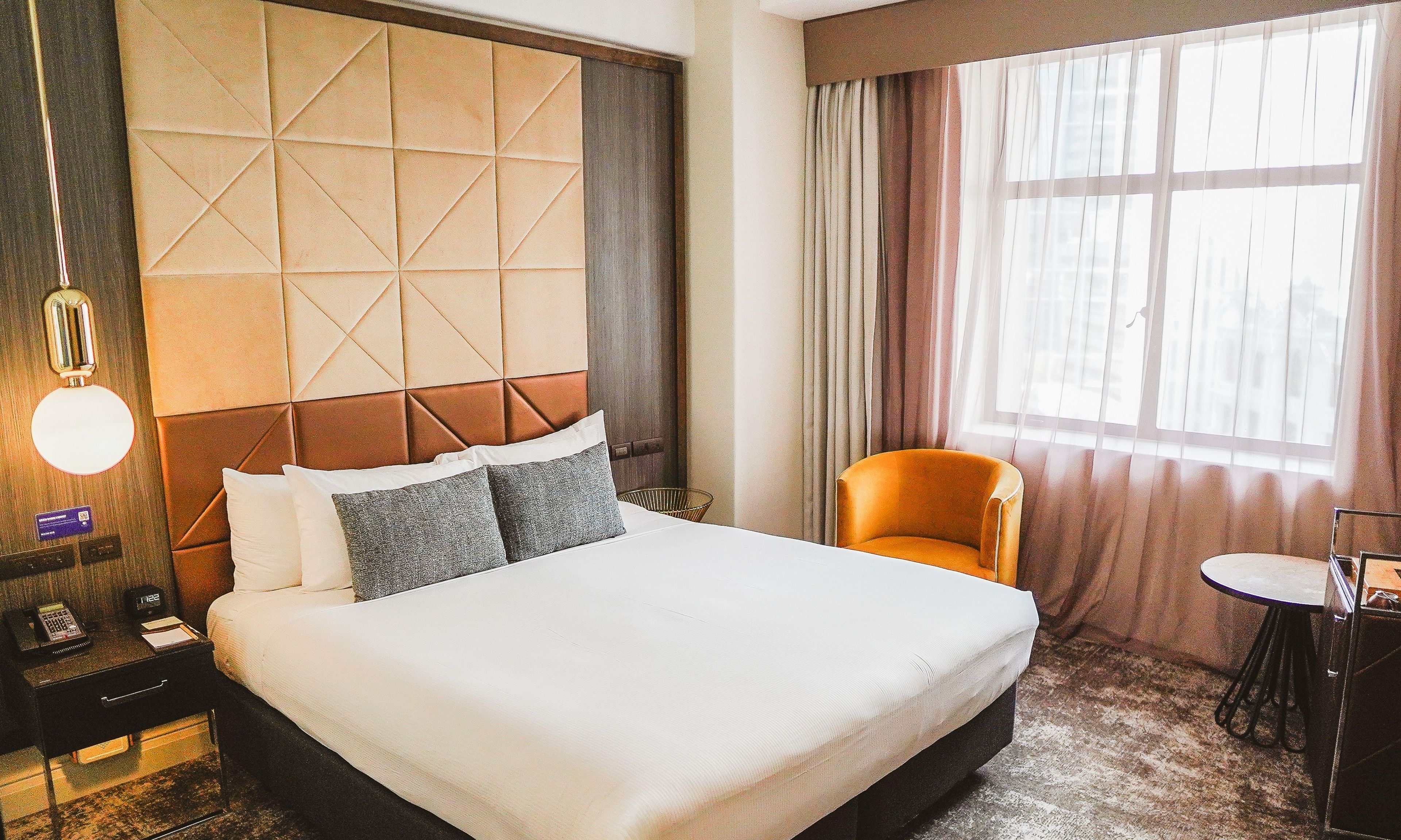 A hotel room with a large bed, a bedside table with a lamp, a chair, a small table, and a window with curtains.