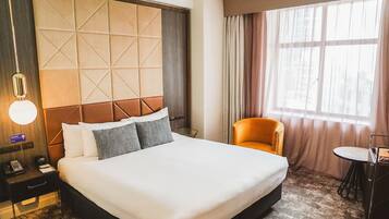 A hotel room with a large bed, a bedside table with a lamp, a chair, a small table, and a window with curtains.