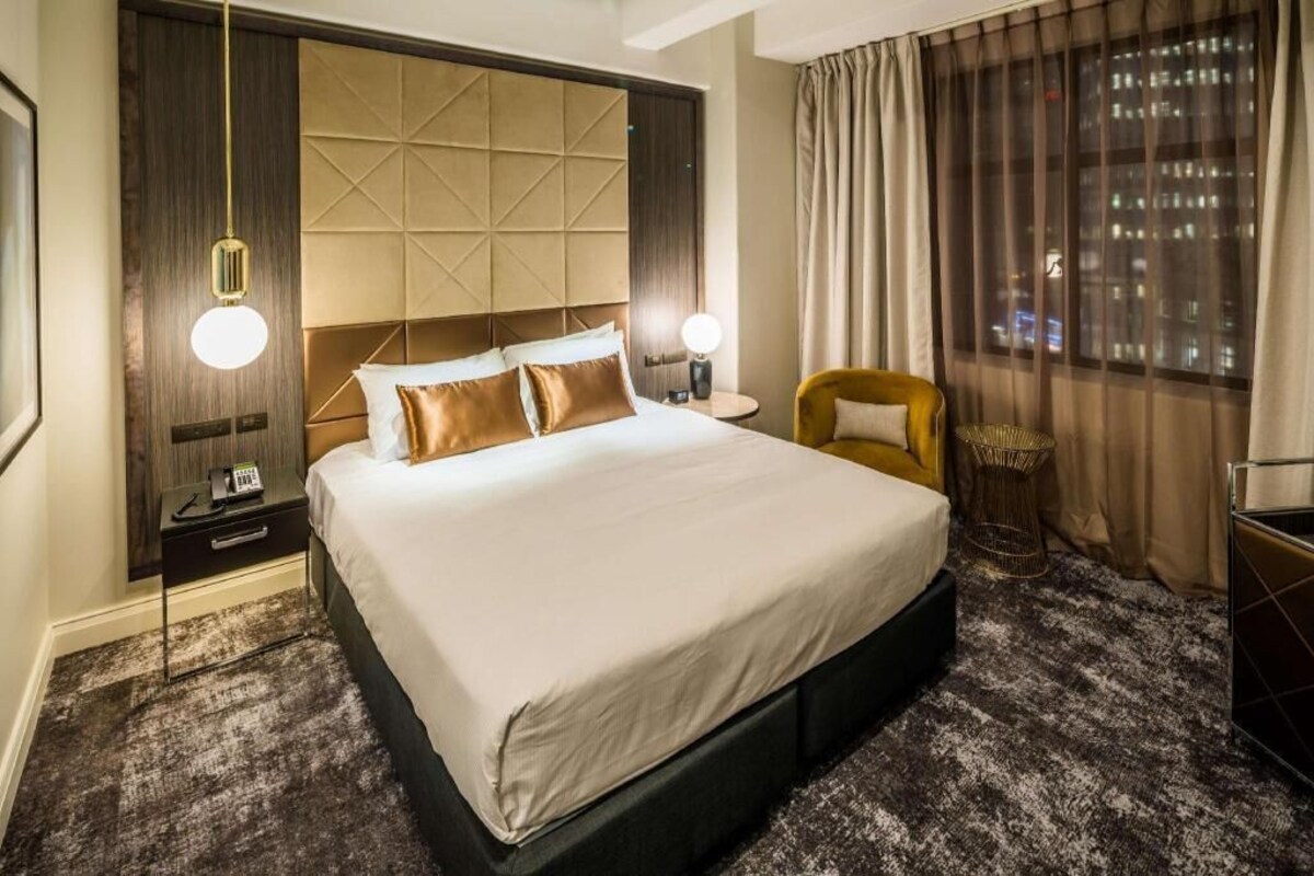 A hotel room with a large bed, bedside lamps, a chair, and a view of the city through the window.