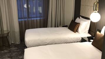 A hotel room with two beds, a large window with curtains, a round light fixture, and a textured wall.