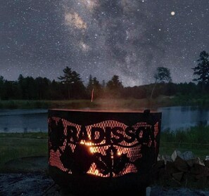 A campfire with a metal grill, surrounded by a natural landscape with trees and a lake under a starry night sky. - Radisson Hotel Kingswood Hotel Fredericton, NB (Hanwell)