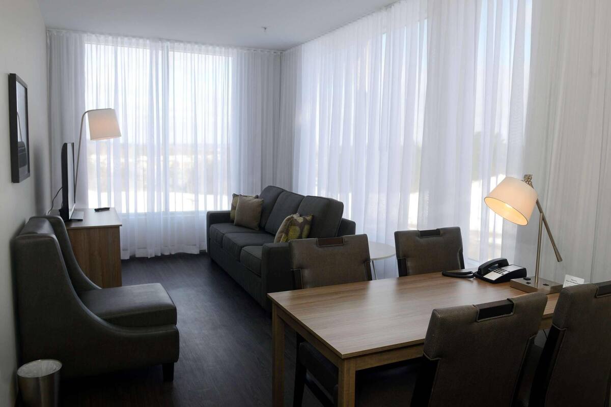 A modern hotel room with a sofa, two armchairs, a wooden table, and a desk with a lamp.