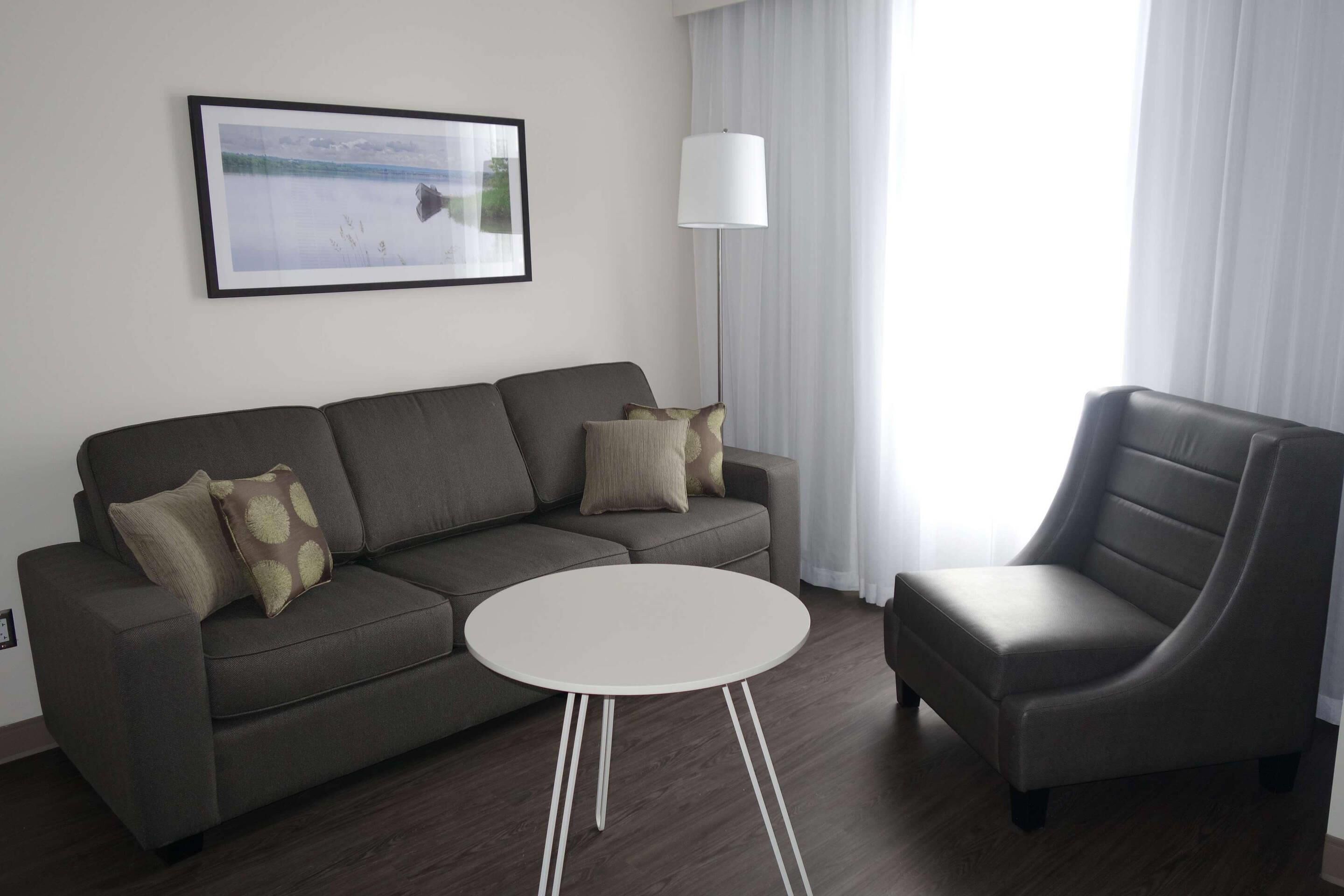 A modern living room with a grey sofa, a round white coffee table, and a grey armchair. A framed picture hangs on the wall, and a floor lamp is positioned near the window.