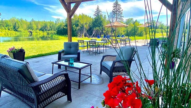 An outdoor patio with wicker furniture, a table, and chairs, overlooking a lake and greenery. - Radisson Hotel Kingswood Hotel Fredericton, NB (Hanwell)