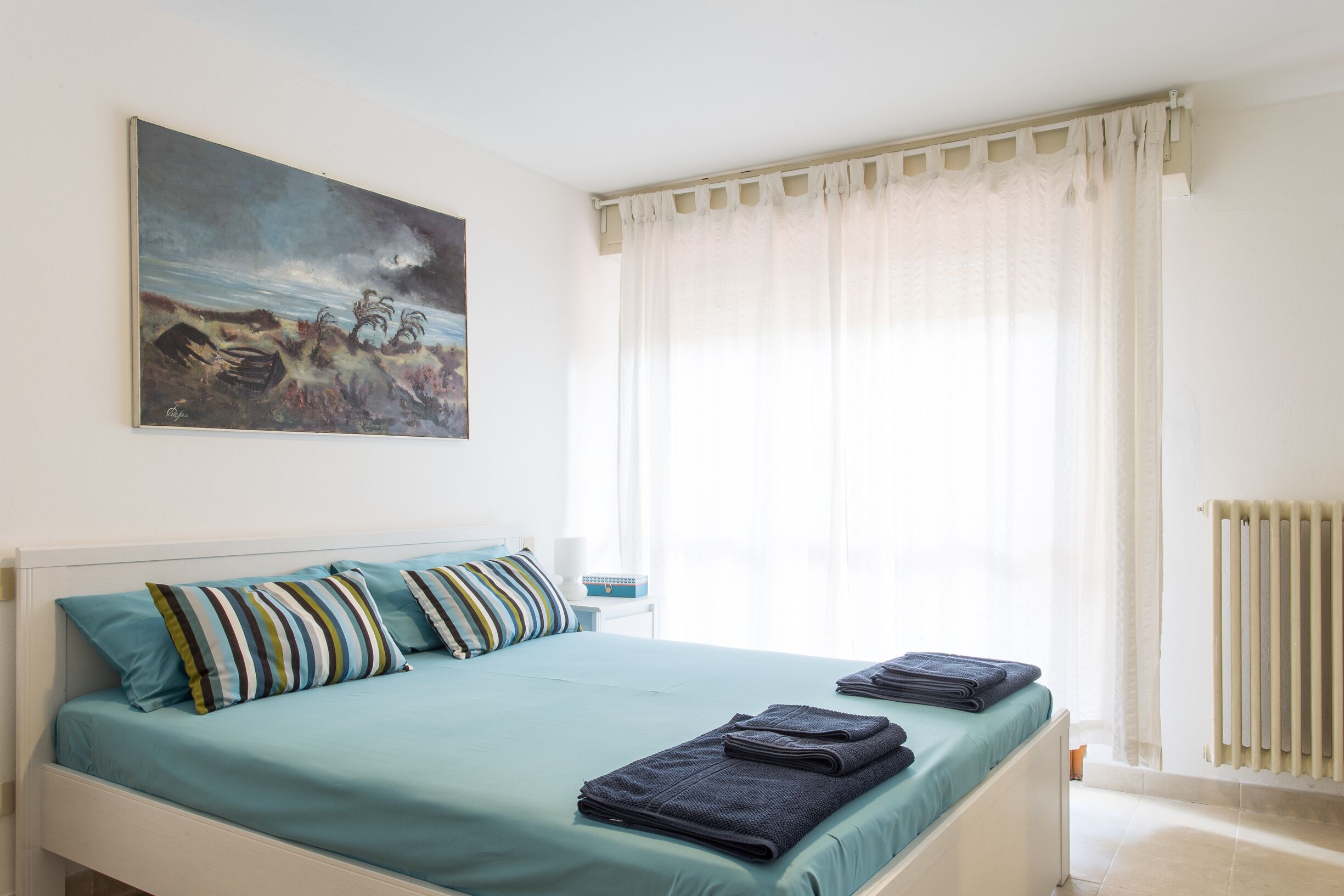 A neatly made bed with striped pillows, a painting on the wall, and a window with sheer curtains.