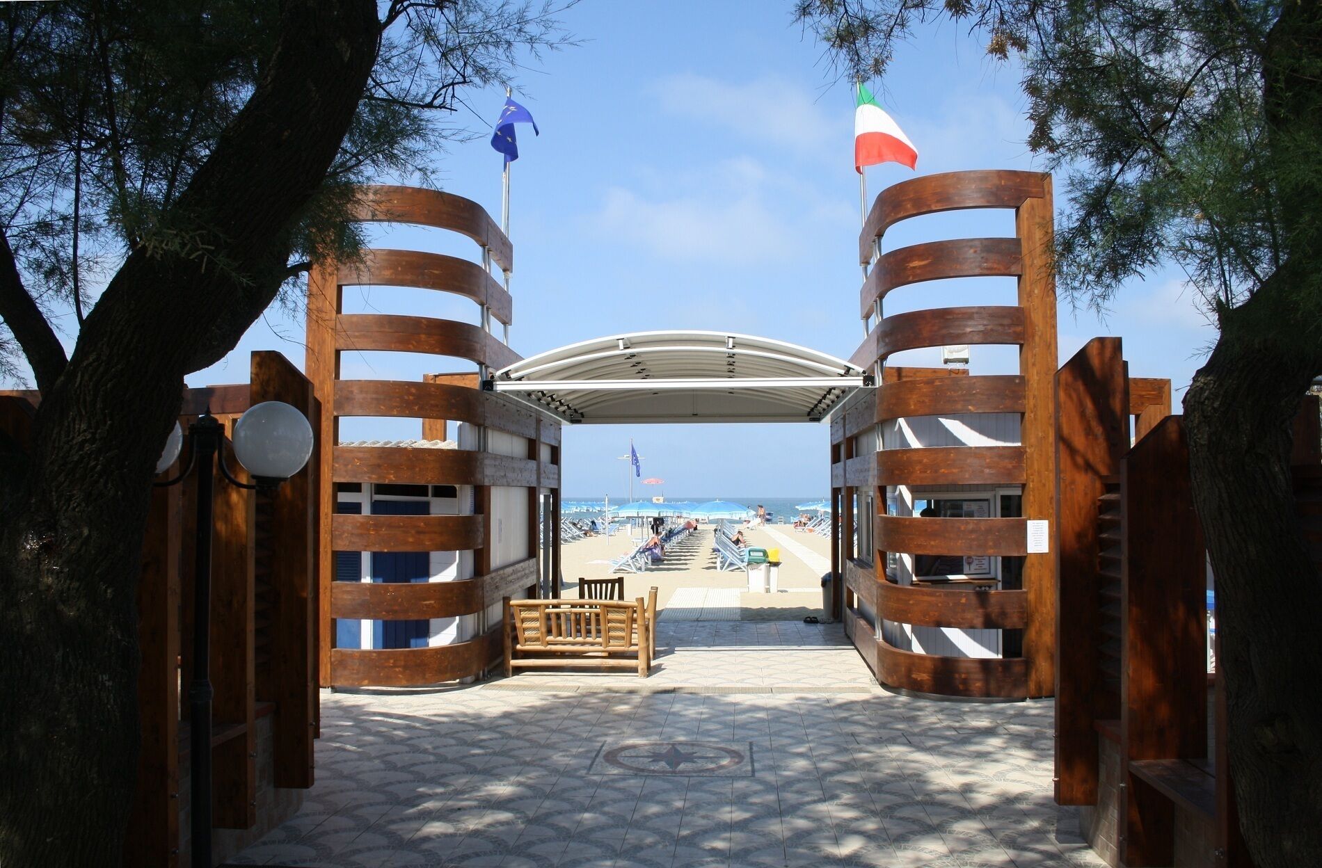 Une porte en bois, surmontée d’un toit incurvé, mène à une zone de plage où se trouve un banc et d’où l’on peut admirer la mer.