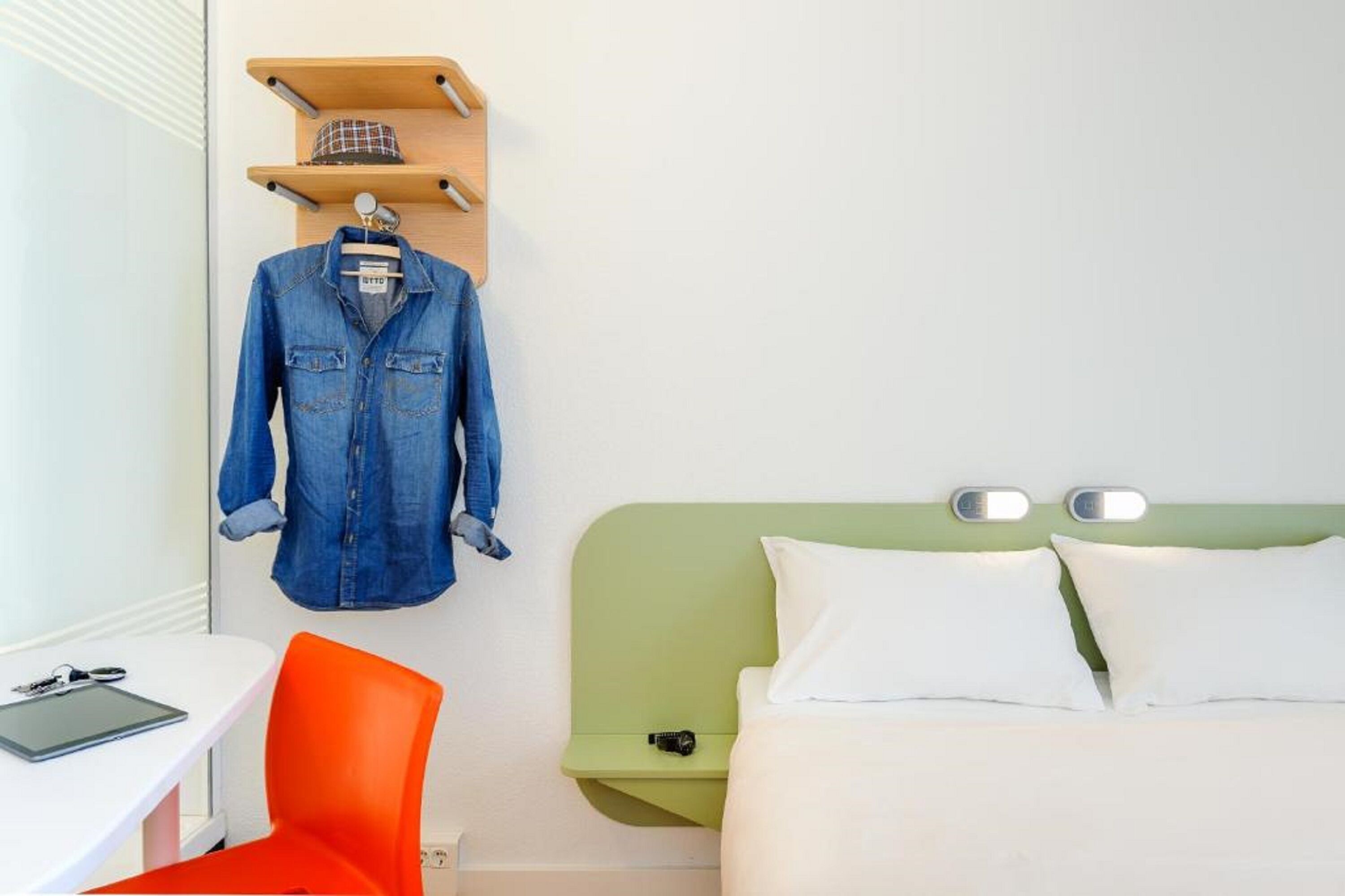 Une chambre d’hôtel avec un lit, un bureau et une chaise, une veste accrochée et une étagère sur laquelle se trouve un chapeau.