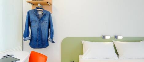 Habitación de hotel con cama, escritorio con silla, una chaqueta colgada y repisa con sombrero.