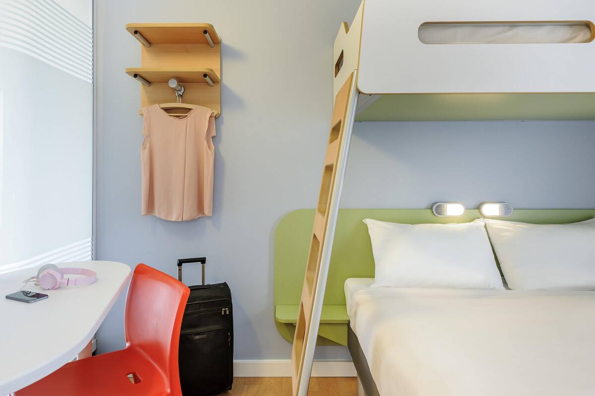 A hotel room with a bunk bed, a desk with a chair, a suitcase, and a hanging coat rack.