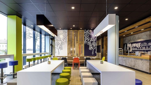 A modern cafe with long communal tables, green and yellow stools, and a counter with a menu. - Ibis Budget Bamberg (Bamberg)