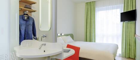 Habitación de hotel con cama, una silla roja, un lavamanos blanco, un espejo y un televisor montado en la pared.