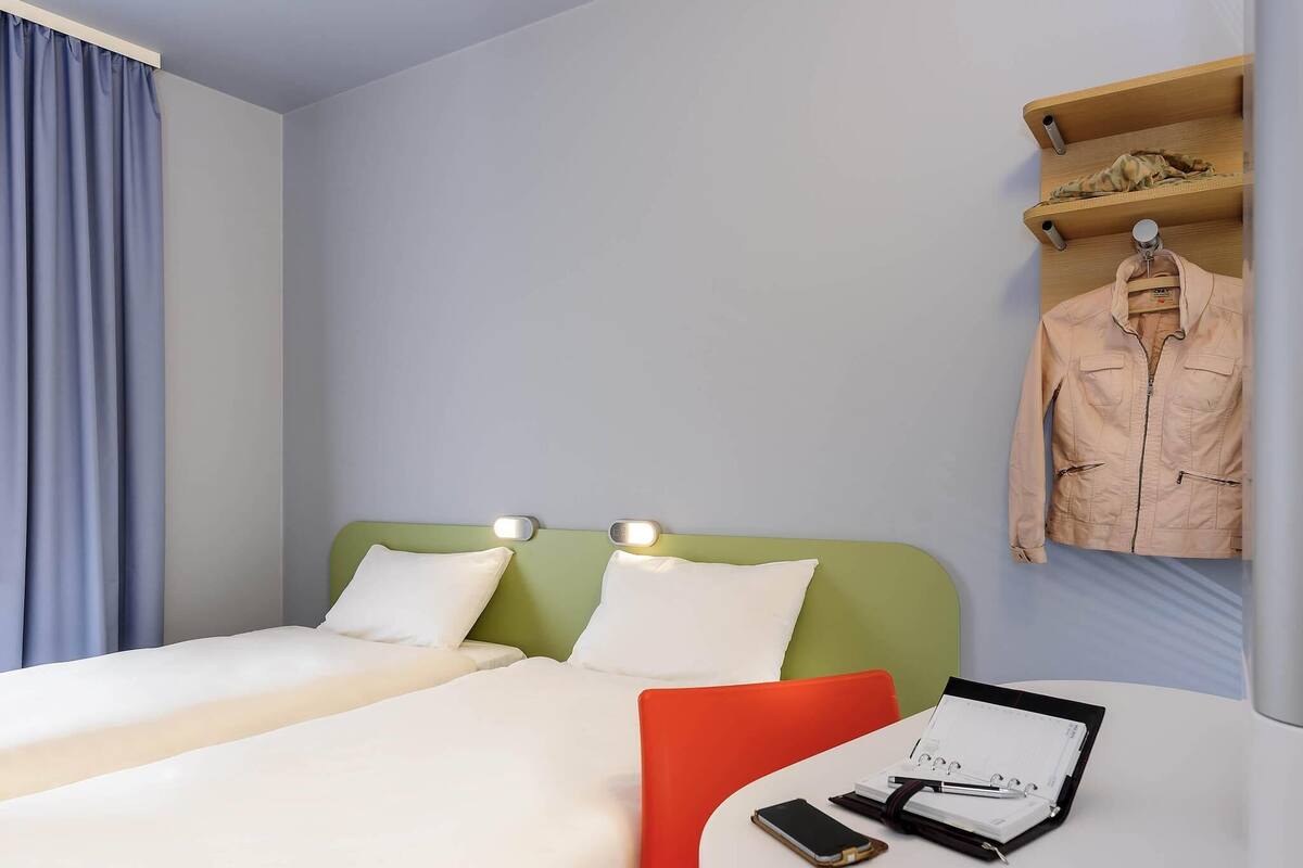 A hotel room with two beds, a small table with a notebook and pen, a smartphone, and a hanging jacket.