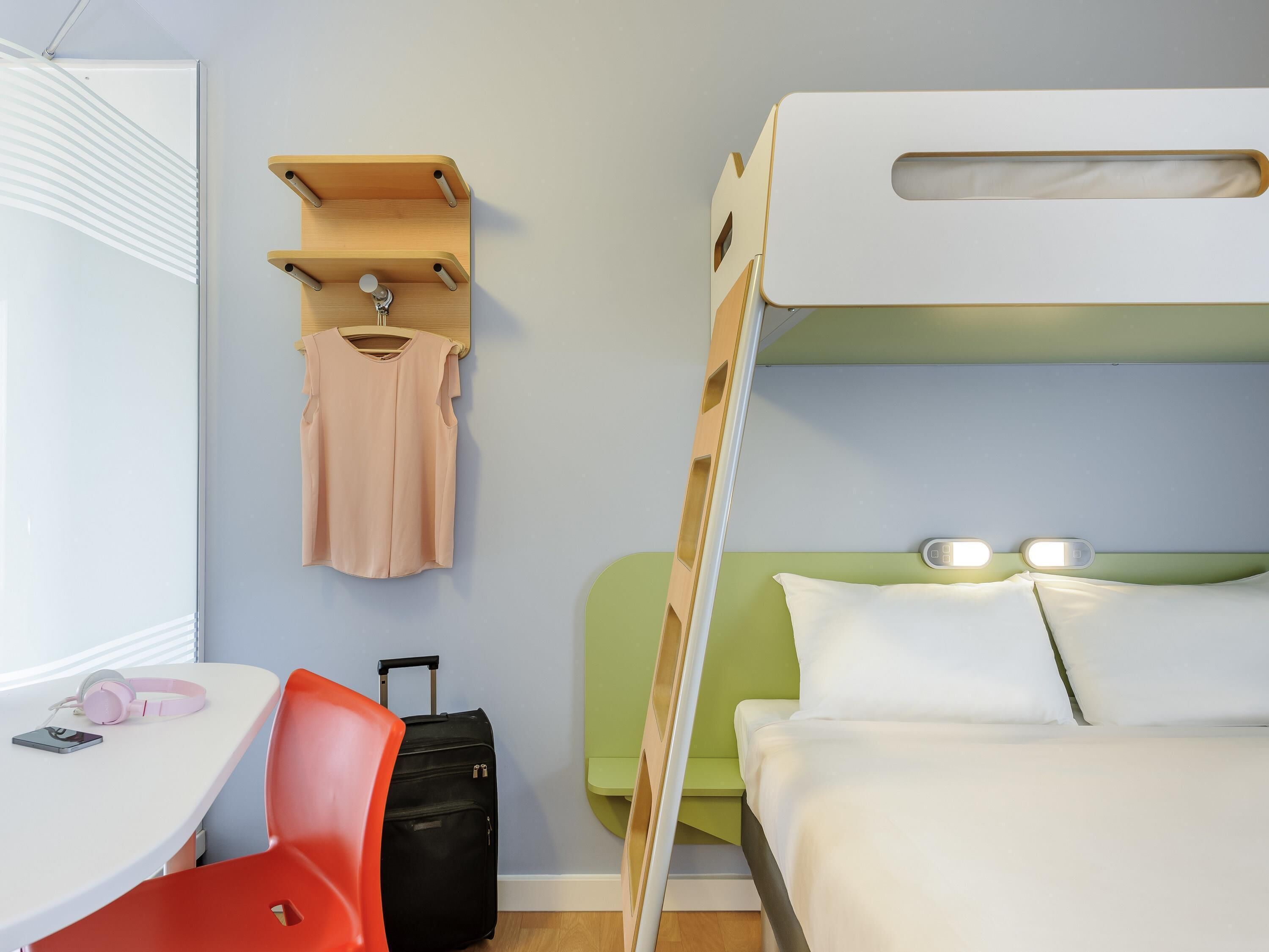 A hotel room with a bunk bed, a desk with a chair, a suitcase, and a hanging coat rack.