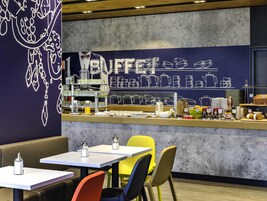 Zona tipo bufé con una barra que exhibe diferentes alimentos y aderezos. Hay mesas y sillas dispuestas para comer.