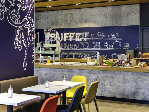 Zona tipo bufé con una barra que exhibe diferentes alimentos y aderezos. Hay mesas y sillas dispuestas para comer.