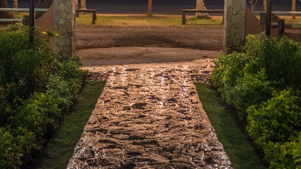 A stone pathway leading to a thatched-roof structure with string lights, surrounded by palm trees and a view of the ocean.