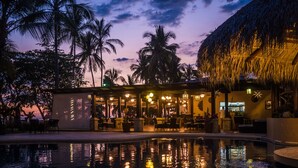 A poolside area with a restaurant, palm trees, and a thatched-roof structure.