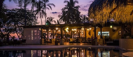 A poolside area with a restaurant, palm trees, and a thatched-roof structure.